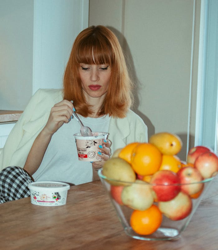 about-img A young woman enjoys yogurt next to a bowl of fresh fruits indoors, blending health and leisure.